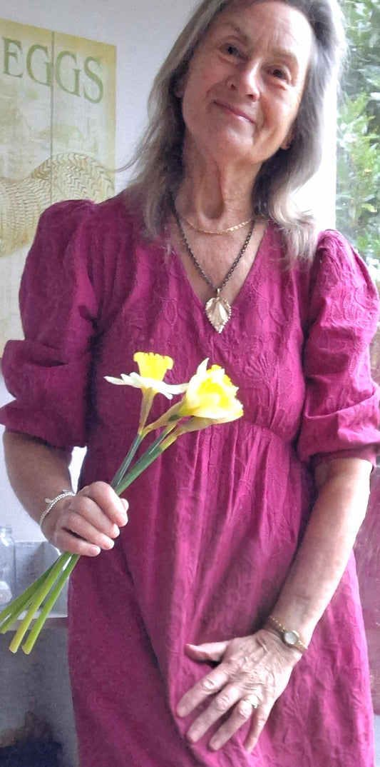 Jane is 67 years young. She wears a  deep pinky/purple linen dress and holds a few fresh daffodils, whilst standing by her patio doors in Cornwall. She has a shiny nettle necklace adorning her decolletage.
